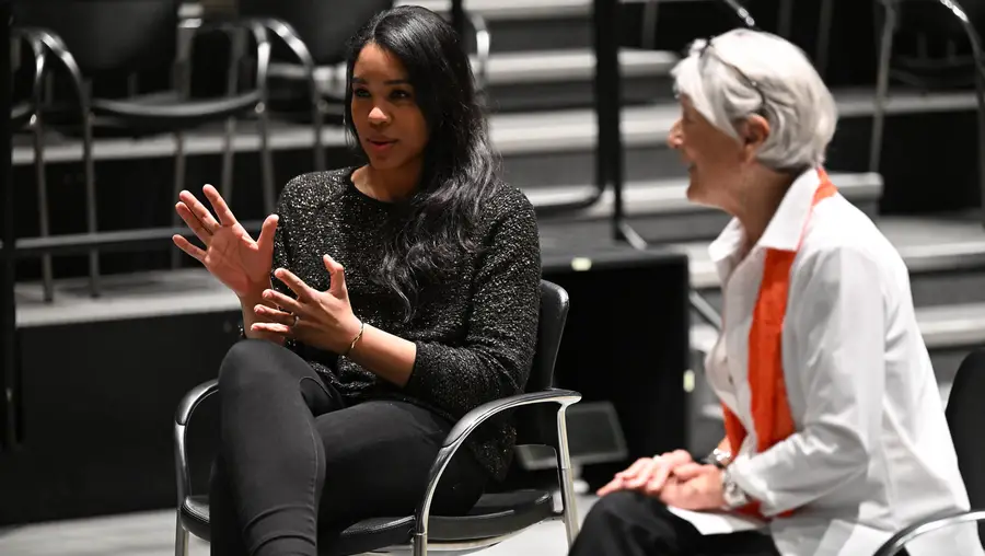 Two women seated in chairs look toward the left and speak to a crowd that is out of frame. The woman on the left is gesturing with her hands.